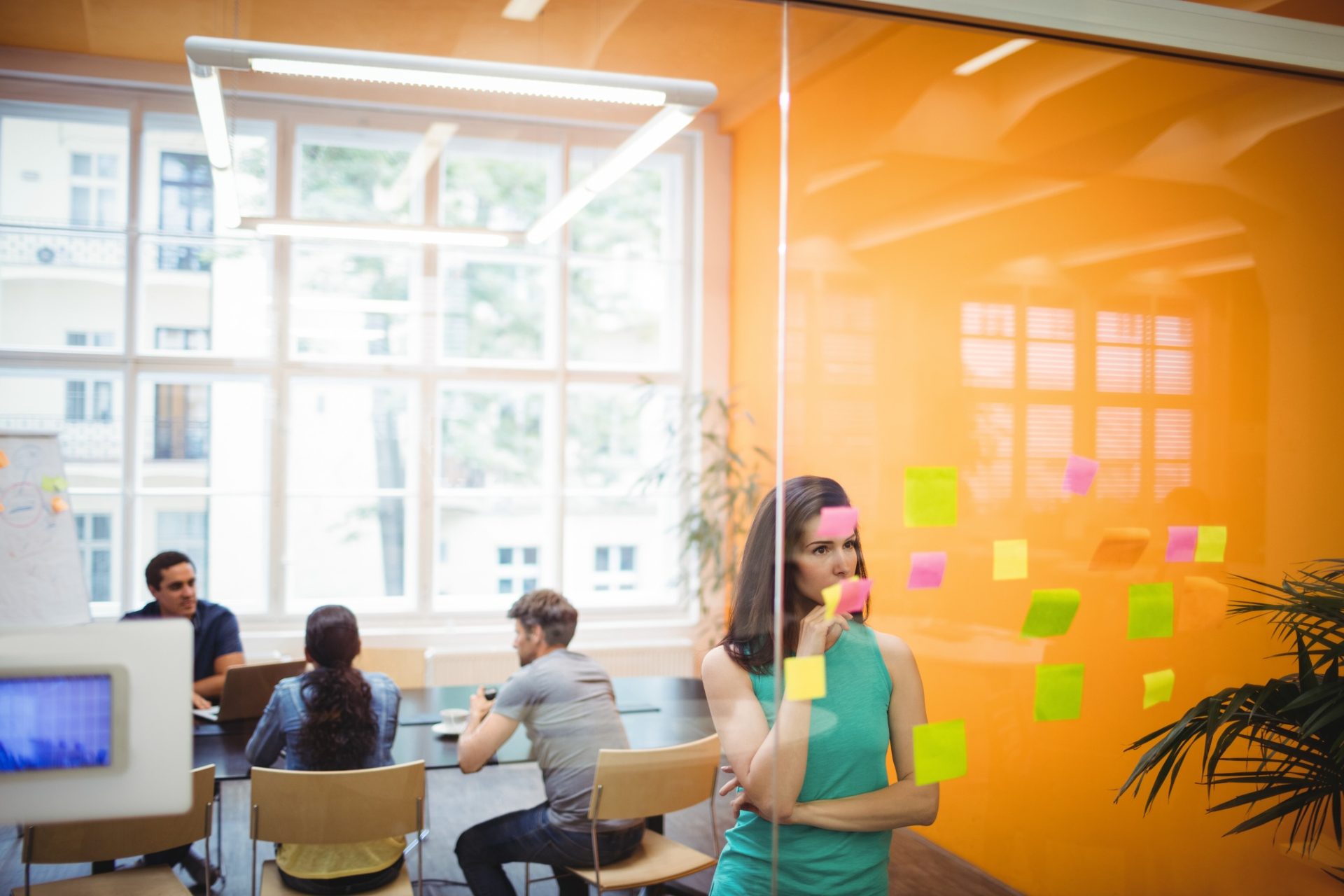 A designer reviewing sticky notes on a glass wall while her team meets in the background